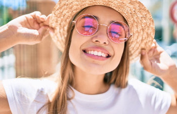 Young woman wearing a straw hat and pink-tinted round sunglasses smiles broadly while holding the brim of her hat.