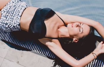 Woman in a black bikini top and patterned bottoms lying on her back by a pool, sunbathing with eyes closed and arms above her head.