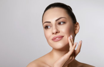 A woman with clear skin and dark hair pulled back touches her face gently while looking slightly to the side against a plain light background.