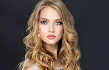 Young woman with long, wavy blonde hair and blue eyes, wearing light makeup, poses against a dark background.