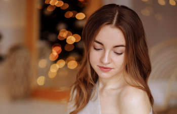 Young woman with long brown hair looking down, wearing an off-shoulder top, with blurred warm lights in the background.