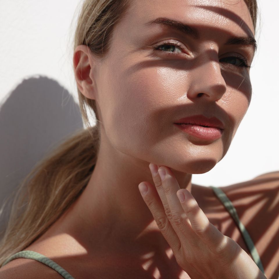 A woman with blonde hair touches her chin while standing in sunlight, with leaf shadows cast across her face and shoulder.